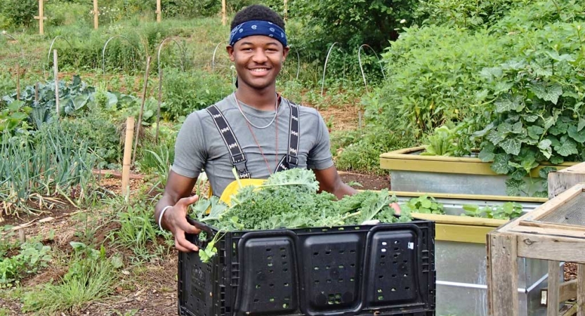 A person smiles at the camera while carrying a box filled with vegetables. A garden is in the background.